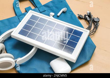 Komposition mit tragbarem Solarpanel, kabellosen Kopfhörern und Tasche auf beigefarbenem Holzhintergrund Stockfoto