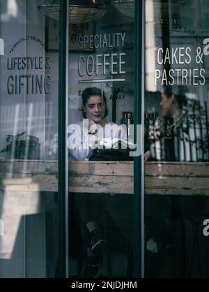 Zwei Frauen teilen sich einen ruhigen Kaffeegenuss am Fenster eines gemütlichen Cafés, umgeben von warmen Holztexturen und handbeschrifteten Schildern mit Spezialitäten Stockfoto