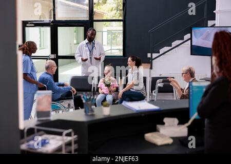 Krankenschwester bringt älteren Mann nach Abschluss der Konsultation, während der Arzt der Familie im Wartebereich des Krankenhauses die medizinische Behandlung erklärt. Ein kleines Mädchen, das Blumen für Großvater hält. Medizindienst Stockfoto