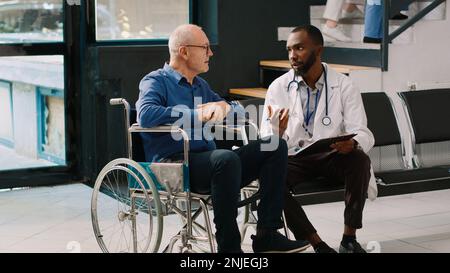 afroamerikanischer Arzt, der Beratung mit dem alten Mann im Rollstuhl, Ausfüllen von medizinischen Berichtspapieren in der Krankenhauslobby. Patient mit chronischer Behinderung im Wartezimmer im Gespräch mit dem Arzt. Stockfoto