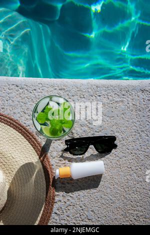 Erfrischender Mojito-Cocktail am Pool mit Sonnencreme, Sonnenbrille und Strohhut Stockfoto