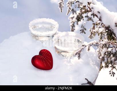Zwei Kristallgläser Sekt oder Champagner und rotes Herz im Schnee auf winterlichem natürlichen Hintergrund mit wunderschönem Sonnenlicht-Bokeh. Liebe und Romantik. Stockfoto