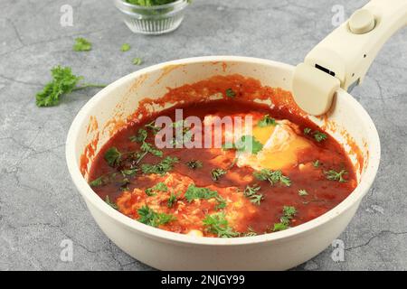 Shakshuka, pochierte Eier in Tomatensoße, Zwiebeln, Pfeffer und Gewürzen auf Weißpfanne auf grauem Zement-Hintergrund. Berühmter traditioneller arabischer und israelischer Kurzurlaub Stockfoto