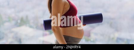 Pränatales Yoga und Bewegung, während eine schwangere Frau zum pränatalen Yoga-Kurs geht und einen wunderschönen Babybauch zeigt, der Yoga-Matte im Profil hält. Eine Frau Stockfoto