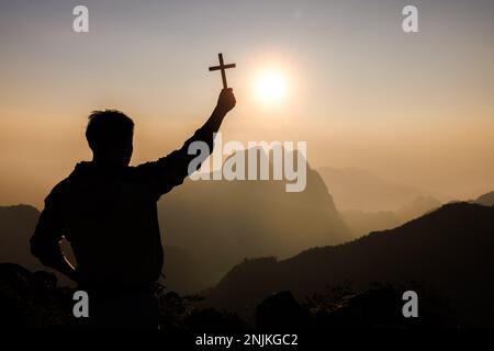 Silhouette eines jungen Mannes, der betet und das christliche Kreuz hält, weil er Gott bei Sonnenuntergang anbetet. Christliche Religion - Vorgeschichte. Stockfoto