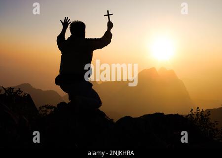 Silhouette eines jungen Mannes, der betet und das christliche Kreuz hält, weil er Gott bei Sonnenuntergang anbetet. Christliche Religion - Vorgeschichte. Stockfoto