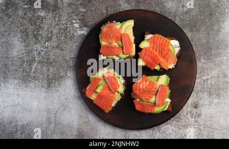Hausgemachte Lachs-Avocado-Sandwiches mit Quark auf einem runden Holzschneidebrett auf dunkelgrauem Hintergrund. Draufsicht, flach liegend Stockfoto