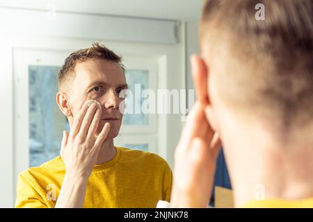 Ein Mann mittleren Alters trägt Gesichtscreme mit einem Wattepad auf und massiert dann mit den Händen. Männliches Porträt im Spiegel. Hautpflege Morgenroutine, sorgfältige Schönheitsbehandlung Stockfoto