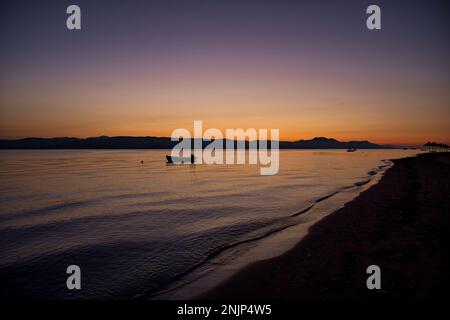 Sonnenaufgang am Meer auf einer Kette von Hügeln, der Himmel scheint in goldorange, violett und blau und spiegelt sich im Wasser, auf dem Meer ein kleines Boot. Stockfoto