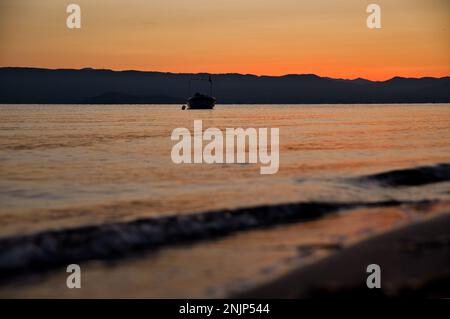 Sonnenaufgang am Meer auf einer Kette von Hügeln, der Himmel scheint in intensivem goldenen Orange und spiegelt sich im Wasser, auf dem Meer ein kleines Boot. Stockfoto