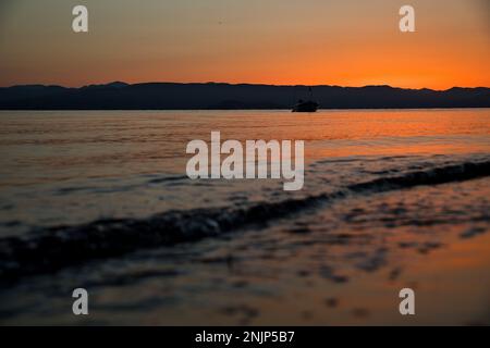 Sonnenaufgang am Meer auf einer Kette von Hügeln, der Himmel scheint in intensivem goldenen Orange und spiegelt sich im Wasser, auf dem Meer ein kleines Boot. Stockfoto