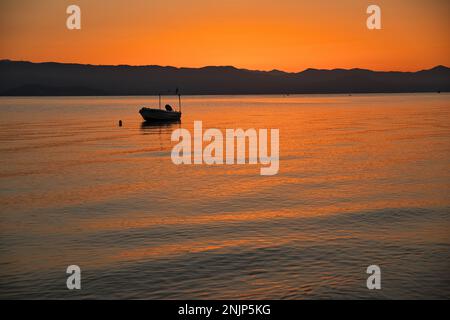 Sonnenaufgang am Meer auf einer Kette von Hügeln, der ganze Himmel scheint in einer intensiven goldenen Orange und spiegelt sich im Wasser, auf dem Meer ein kleines Boot. Stockfoto