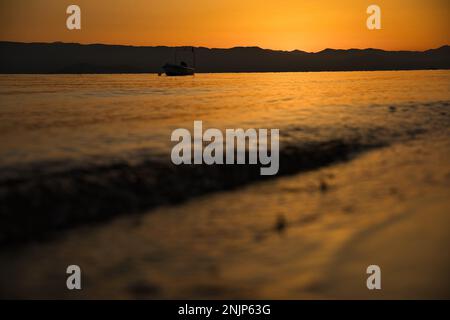 Sonnenuntergang über dem Meer auf einer Kette von Hügeln, der ganze Himmel scheint in goldgelben Farben, die Sonne spiegelt sich im Wasser, auf dem Meer ein kleines Boot. Stockfoto