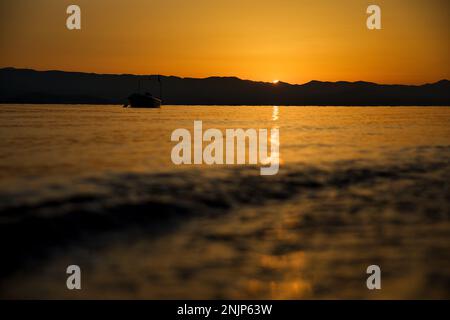 Sonnenuntergang über dem Meer auf einer Kette von Hügeln, der ganze Himmel scheint in goldgelben Farben, die Sonne spiegelt sich im Wasser, auf dem Meer ein kleines Boot. Stockfoto