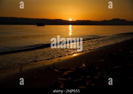 Sonnenuntergang über dem Meer auf einer Kette von Hügeln, der ganze Himmel scheint in goldgelben Farben, die Sonne spiegelt sich im Wasser, auf dem Meer ein kleines Boot. Stockfoto