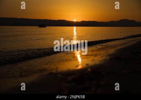 Sonnenuntergang über dem Meer auf einer Kette von Hügeln, der ganze Himmel scheint in goldgelben Farben, die Sonne spiegelt sich im Wasser, auf dem Meer ein kleines Boot. Stockfoto