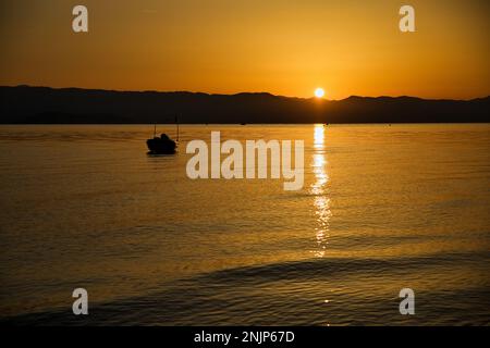 Sonnenuntergang über dem Meer auf einer Kette von Hügeln, der ganze Himmel scheint in goldgelben Farben, die Sonne spiegelt sich im Wasser, auf dem Meer ein kleines Boot. Stockfoto
