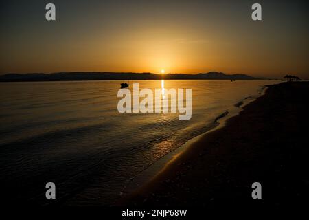 Sonnenuntergang über dem Meer auf einer Kette von Hügeln, der ganze Himmel scheint in goldgelben Farben, die Sonne spiegelt sich im Wasser, auf dem Meer ein kleines Boot. Stockfoto
