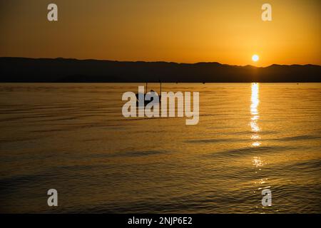 Sonnenuntergang über dem Meer auf einer Kette von Hügeln, der ganze Himmel scheint in goldgelben Farben, die Sonne spiegelt sich im Wasser, auf dem Meer ein kleines Boot. Stockfoto