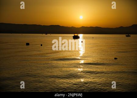 Sonnenuntergang über dem Meer auf einer Kette von Hügeln, der ganze Himmel scheint in goldgelben Farben, die Sonne spiegelt sich im Wasser, auf dem Meer ein kleines Boot. Stockfoto