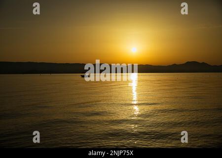 Sonnenuntergang über dem Meer auf einer Kette von Hügeln, der ganze Himmel scheint in goldgelben Farben, die Sonne spiegelt sich im Wasser, auf dem Meer ein kleines Boot. Stockfoto
