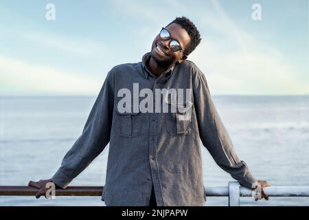 Ein junger erwachsener afrikanischer Mann mit freundlichem Ausdruck, der sich mit dem Meer im Hintergrund auf ein Geländer lehnt. Er trägt ein graues Hemd und eine verspiegelte Sonnenbrille Stockfoto