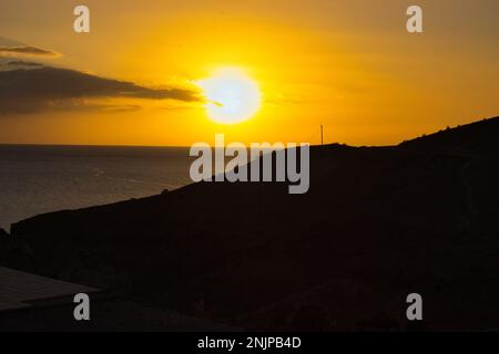 Sonnenuntergang am Meer, die Sonne nahe am Horizont, der ganze wolkige Himmel scheint in intensiven goldenen Farben, im Vordergrund ein Hügel im Schatten. Stockfoto