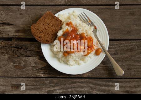 Weißer Teller mit gekochtem Reis und Bohnen mit Brot auf einem alten Holzbrett Stockfoto