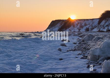 Sonnenuntergang an einem verschneiten Strand hinter einem Hügel, der Himmel scheint in goldenen und pfirsichfarbenen Orange Farben. Stockfoto