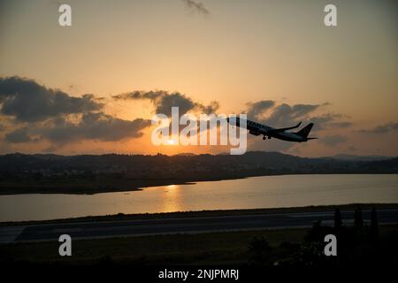 Sonnenuntergang am Meer, die Sonne auf einer Kette von Hügeln, der wolkige Himmel scheint in goldorangefarbenen Farben, im Vordergrund ein fliegendes Flugzeug. Stockfoto