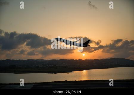 Sonnenuntergang am Meer, die Sonne auf einer Kette von Hügeln, der wolkige Himmel scheint in goldorangefarbenen Farben, im Vordergrund ein fliegendes Flugzeug. Stockfoto