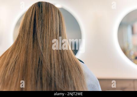 Niedrige Rückansicht einer anonymen Kundin, die in der Nähe eines runden Spiegels sitzt, mit Beleuchtung im modernen Kosmetikstudio Stockfoto