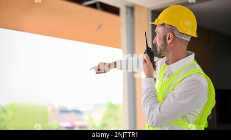 Seriöser und entschlossener weißer Vorarbeiter oder Bauleiter in einem gelben Schutzhelm, der ein Walkie-Talkie benutzt, um seine Bauarbeiter zu befehligen. B Stockfoto