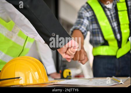 Gekürzt und Nahaufnahme eines Teams von Bauarbeitern, das mit einem Geschäftsmann auf der Baustelle Faustschläge macht. Teamwork-Konzept Stockfoto