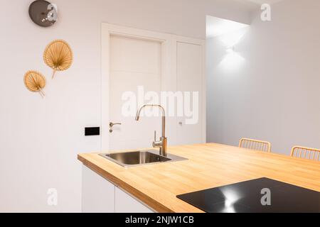 Nahaufnahme der Kücheninsel. Tischplatte aus Holz in einer honigfarbenen Farbpalette. Die Arbeitsfläche verfügt über ein eingebautes Spülbecken und eine Keramikplatte, über der sich die Abluftanlage befindet Stockfoto