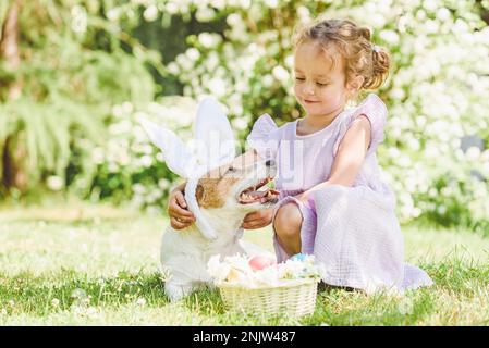 Süßes kleines Mädchen, das ihren Hund umarmt und Osterhäschen-Ohren-Kostüm trägt. Korb mit dekorierten Eiern und Blumen. Stockfoto