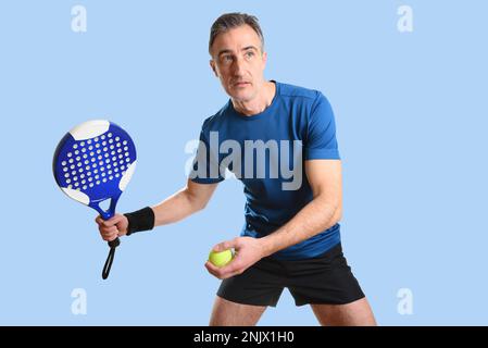 Porträt eines Mannes, der Padel in Position spielt, um zu dienen, mit Schläger und Ball in den Händen und blau-schwarzem Sportanzug auf blauem, isoliertem Hintergrund. Front V Stockfoto