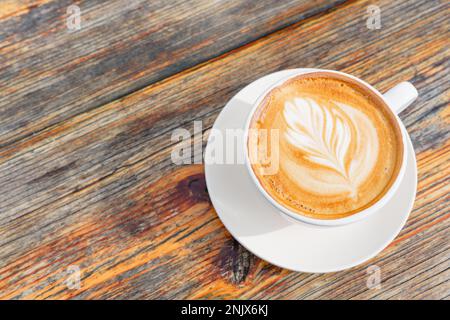 Perfekt gegossener Latte, komplett mit aufwändiger Latte Art, auf einer weißen Untertasse auf einem rustikalen Holztisch. Die Schönheit einer gut gemachten Tasse Kaffee Stockfoto