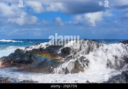 Raues Gelände an der Küste Hawaiis, während Wellen gegen die felsige Oberfläche prallen und Wasser in die Luft spritzt, während der Regenbogen in die Luft spritzt. Stockfoto