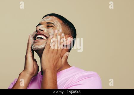 Ein Mann lächelt fröhlich, während er ein Gesichtswaschmittel in einem Studio aufträgt und seine Haut mit dem Reinigungsmittel sanft massiert. Ein hübscher junger Mann, der seine Skinca startet Stockfoto