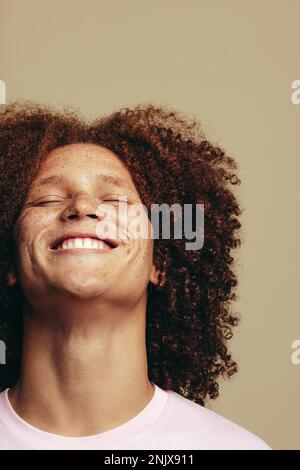 Ein junger Mann mit Sommersprossen und gelocktem Ingwerhaar lächelt mit geschlossenen Augen in einem Studio und strahlt Selbstliebe und Selbstvertrauen aus. Glücklicher junger Mann umarmt Stockfoto