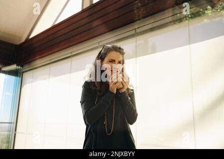 Fröhliche Seniorin lächelt, während sie eine Tasse Tee in ihrem Wohnzimmer trinkt. Glückliche reife Frau, die einen entspannten Morgen zu Hause genießt. Stockfoto