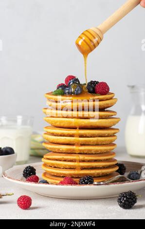 Köstliches und herzhaftes Frühstück - üppige Pfannkuchen mit Beeren über Honig auf grauem Teller, vertikales Bild Stockfoto