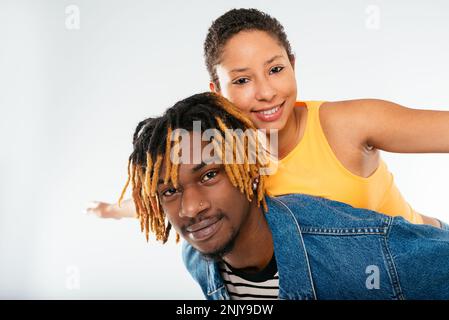 Positiver afroamerikanischer Freund mit gefärbtem Haar in Dreadlocks in Jeansjacke, der einer lächelnden Freundin mit gelbem Oberteil einen Huckepack verpasst Stockfoto