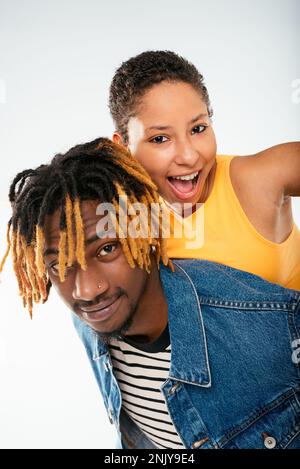 Positiver afroamerikanischer Freund mit gefärbtem Haar in Dreadlocks in Jeansjacke, der einer lächelnden Freundin mit gelbem Oberteil einen Huckepack verpasst Stockfoto
