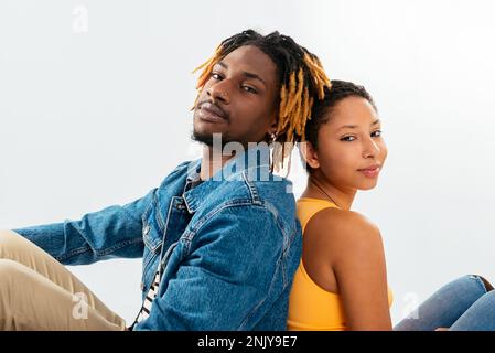 Von oben: Ethnischer Freund und Freundin in Jeansbekleidung, die sich Rücken an Rücken lehnen und sich den Kopf auf die Schultern legen und in die Kamera schauen Stockfoto