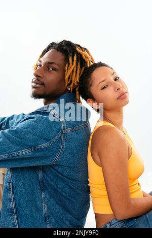 Von oben: Ethnischer Freund und Freundin in Jeansbekleidung, die sich Rücken an Rücken lehnen und sich den Kopf auf die Schultern legen und in die Kamera schauen Stockfoto