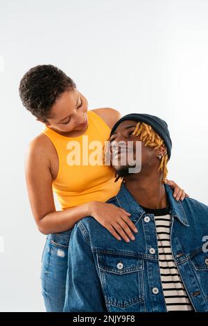 Positive gemischte Rasse Frau mit kurzen Haaren in Jeans und orangefarbenem Oberteil, die sich auf den Rücken eines Mannes in Jeansjacke lehnt, auf weißem Hintergrund und in die Kamera schaut Stockfoto