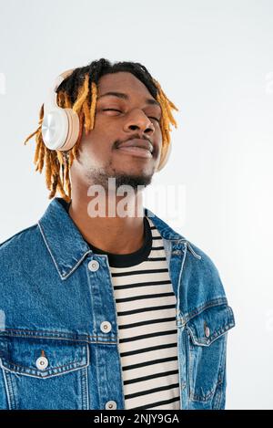 Positiver junger afroamerikanischer Mann mit Bart und Dreadlocks in Jeansjacke, der Musik in kabellosen Kopfhörern auf weißem Hintergrund genießt Stockfoto