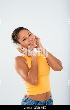 Positives junges weibliches Modell in legerem Outfit, das lächelt, während es Musik in kabellosen Kopfhörern hört und vor weißem Hintergrund auf die Kamera schaut Stockfoto